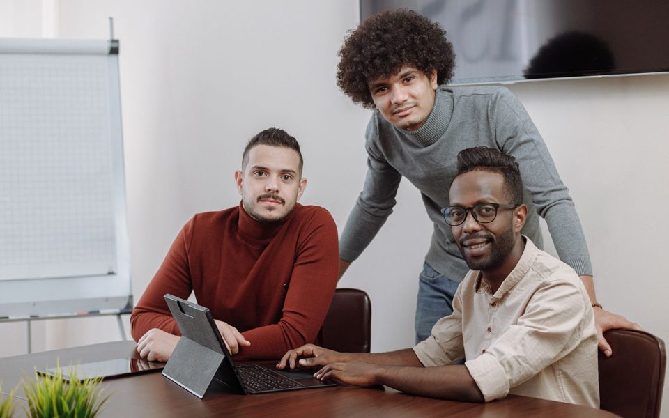Three workplace colleagues gathered around a laptop at a shared desk.